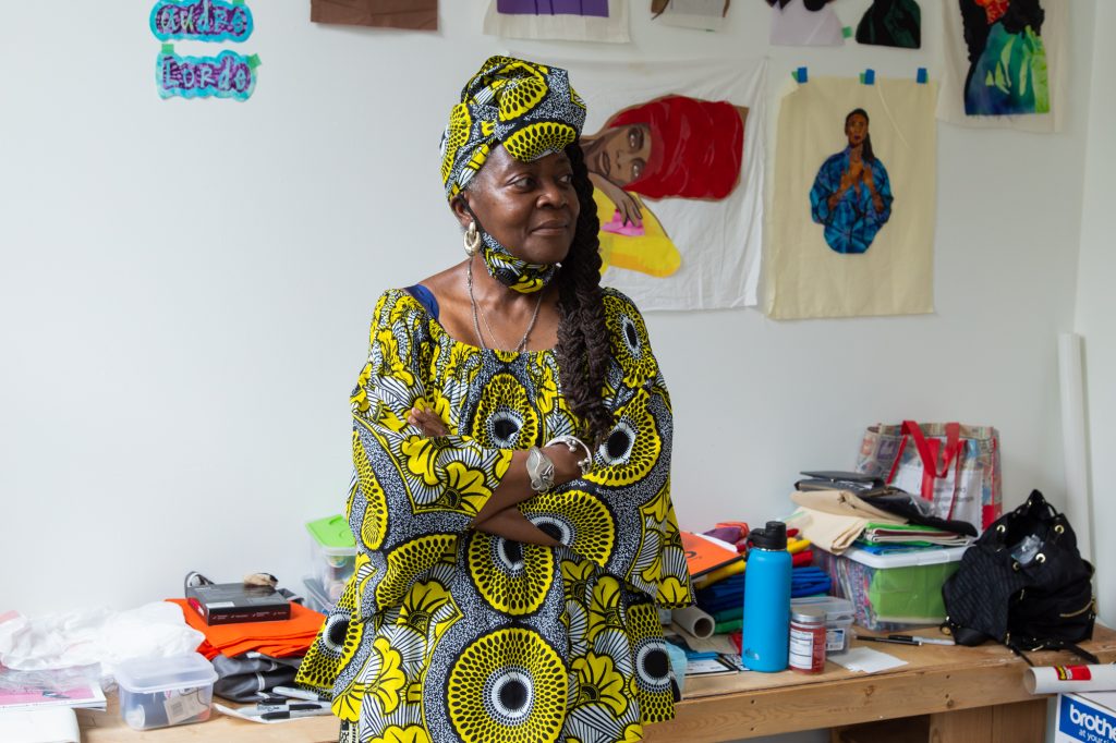 A portrait of artist Dorothy Burge standing with hands folded and looking beyond the frame while in her studio at Hyde Park Art Center. She wears a top, head wrap and pants with black, yellow, and white patterns. Behind her is a bench with sewing and other materials. Along the wall are fabric portraits of iconic Black women, with a portrait of Erykah Badu and the words "Audre Lorde" on the wall in the background. Photo by Joshua Clay Johnson.