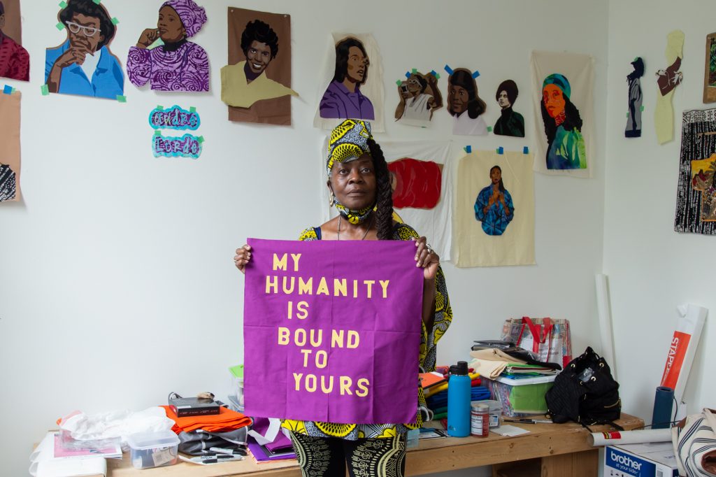 A portrait of Dorothy Burge standing in her studio at Hyde Park Art Center holding a magenta-colored fabric pattern with yellow lettering that reads, "My Humanity is Bound To Yours." Behind her is a bench with sewing and other materials. Along the wall are fabric portraits of iconic Black women. Photo by Joshua Clay Johnson.