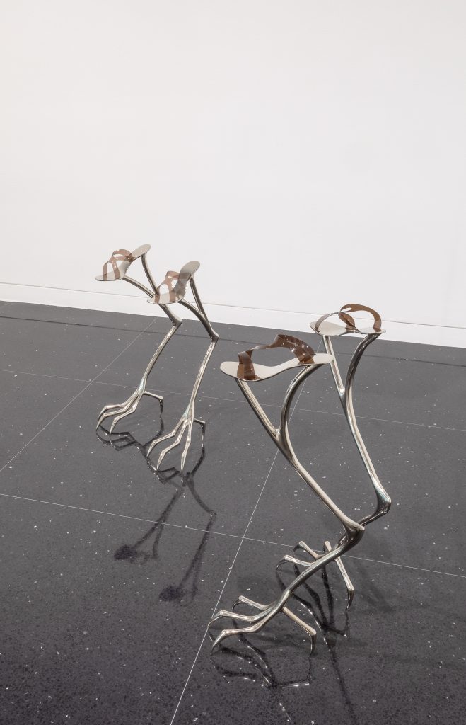 Image: Hannah Levy, "Untitled," 2021. Nickel-plated steel, silicone. 27.75 x 8 x 15 in. & 33.5 x 8 x 15 in. An installation view of two steel sculptures that look similar to ostrich legs. At the top of each of the four legs sits a sandal-like detail. Image courtesy of the Arts Club Chicago.
