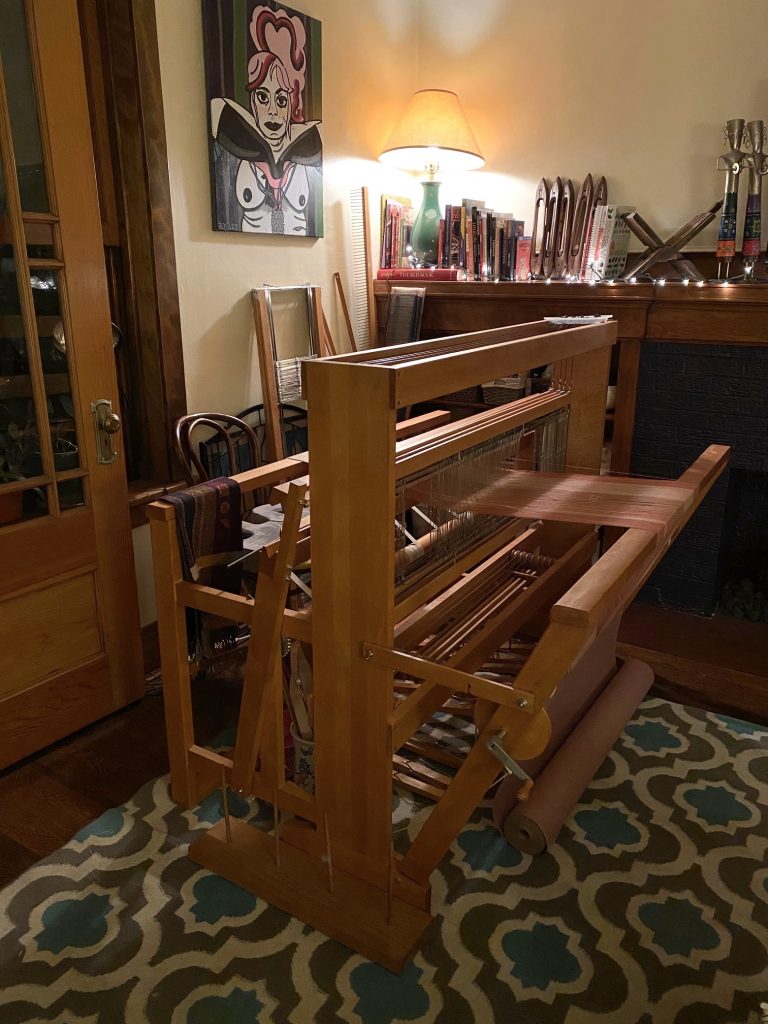 Image: A photo of Sangi Ravichandran's loom in her living space. There are various books and tools in the background and a painting hangs on the wall. Photo courtesy of the artist.