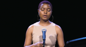 Image: India Sada reads from her poem "She Dreamed of Being Saved" She stands in a dark room and is lit from an overhead soft-blue light. Her eyes are closed. She speaks into a mic.