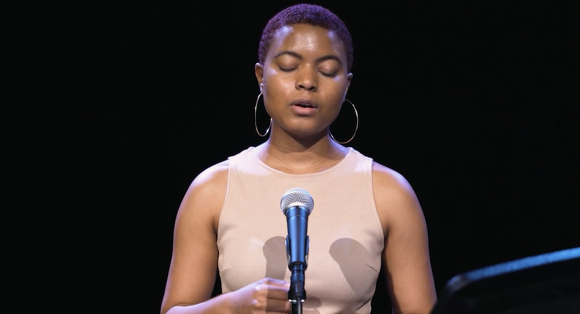 Image: India Sada reads from her poem "She Dreamed of Being Saved" She stands in a dark room and is lit from an overhead soft-blue light. Her eyes are closed. She speaks into a mic.