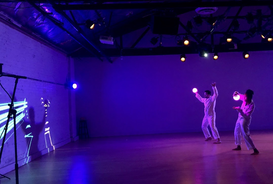 Selena Lasley and Christopher Knowlton, members of Team A, wear white coveralls. Each person holds an orb that is lighting up in their right hand. The orbs are attached to a machine that tracks their movements as lights projected onto a wall, which they both face. There is a microphone stand in the foreground. Photo by Links Hall.
