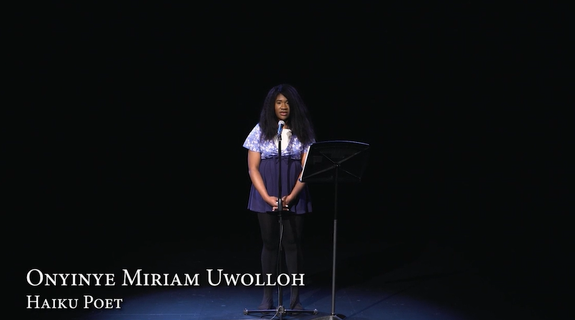 Image: Onyinye Miriam Uwollo reads from her poem "The End Began When". She's in a dark room and is lit from an overhead soft-blue light. Her eyes are closed. She speaks into a mic. Her poem sits on a lectern stand. Image courtesy of Mourning the Creation of Racial Categories Project.