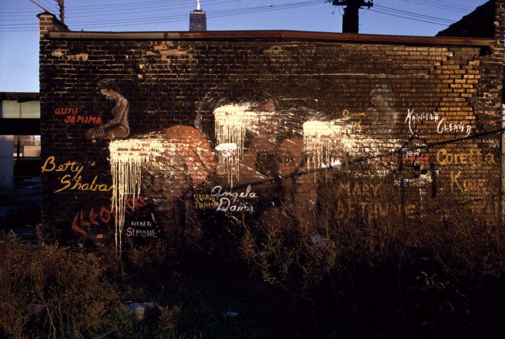 Image: An industrial brick building is in the foreground, painted on it is Vanity Green's mural. Vanita Green, Black Women / Racism, 1970. Mural at 861 N. Orleans. Photograph by Georg Stahl. Courtesy of the Georg Stahl Mural Archive, University of Chicago Visual Resources Center Luna Collection.