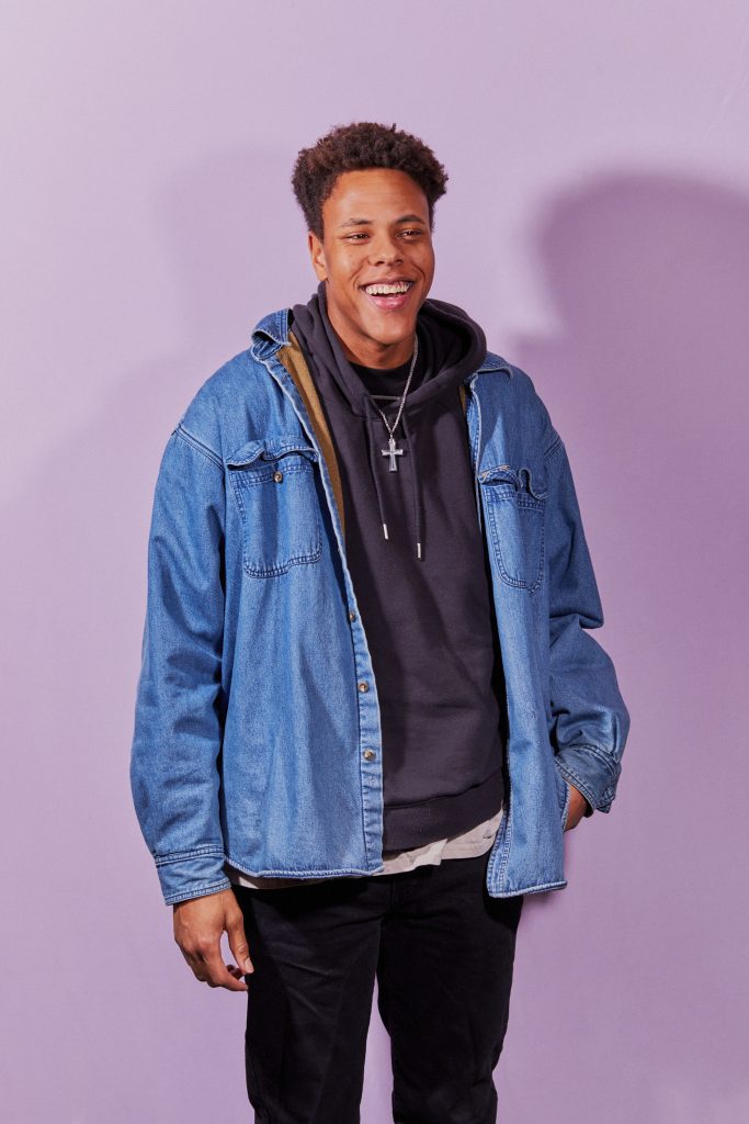 Image: Marcus Pruitt stands with one hand in his pocket in front of a lavender wall. He is wearing a cross necklace, black pants, a black sweatshirt, and a jean jacket. He smiles slightly to the viewer's right. Photo by sarah joyce.