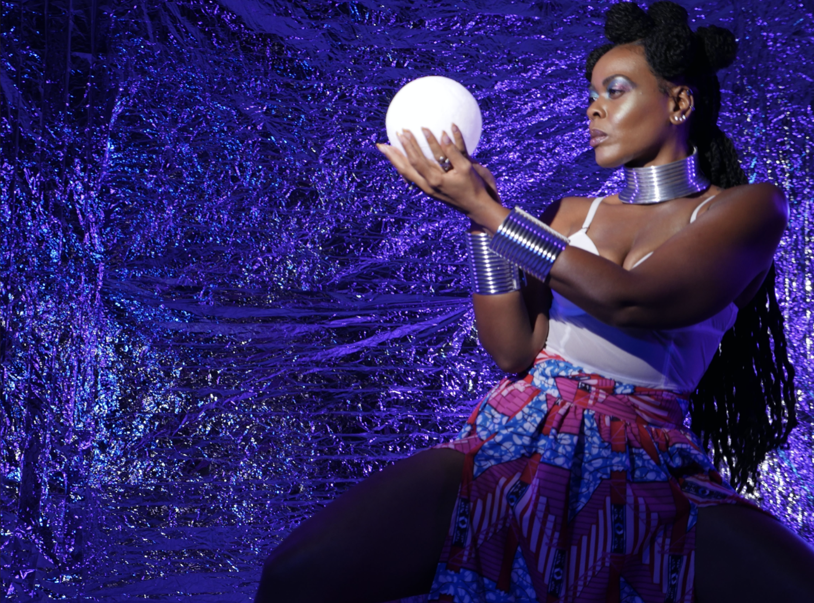 Featured image: A still from, "God is a Black Woman" by Chaz Shermil Hodges. A Black woman holds a white crystal ball against a sparkly purple back drop. She is wearing a blue and pink skirt, a white bustier and silver-ringed bracelets and necklace, suggesting a traditional African attire.