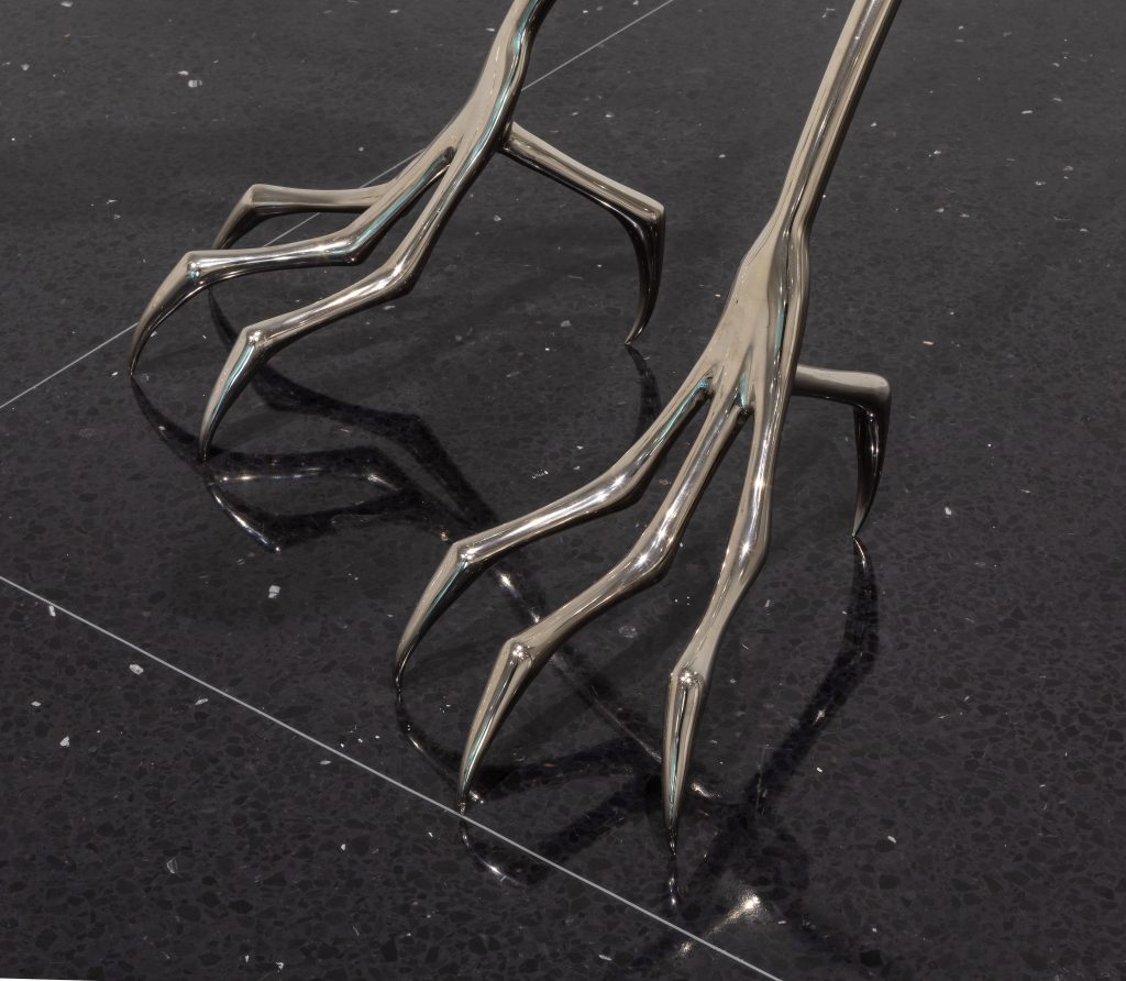 Image: A detail shot of Hannah Levy's sculpture "Untitled," 2021. Nickel-plated steel, silicone. 27.75 x 8 x 15 in. The shot shows a close-up view of two steel ostrich-like feet standing on a black, reflective floor. Image courtesy of the Arts Club Chicago.