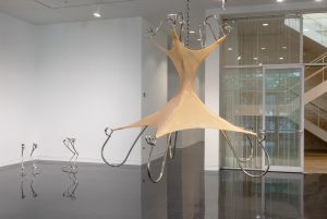 Featured image: An installation view of Hannah Levy's piece "Untitled," 2021. Nickel-plated steel, silicone. 68 x 74 x 74 in. A chandelier-like sculpture that takes the shape of an exaggerated bodice. The bodice-shape is beige with a corset-like detail. Steel, arm-like, structures appear to be holding the shape in place. Another smaller steel sculpture by Hannah Levy can be seen in the background on the left. Image courtesy of the Arts Club Chicago.