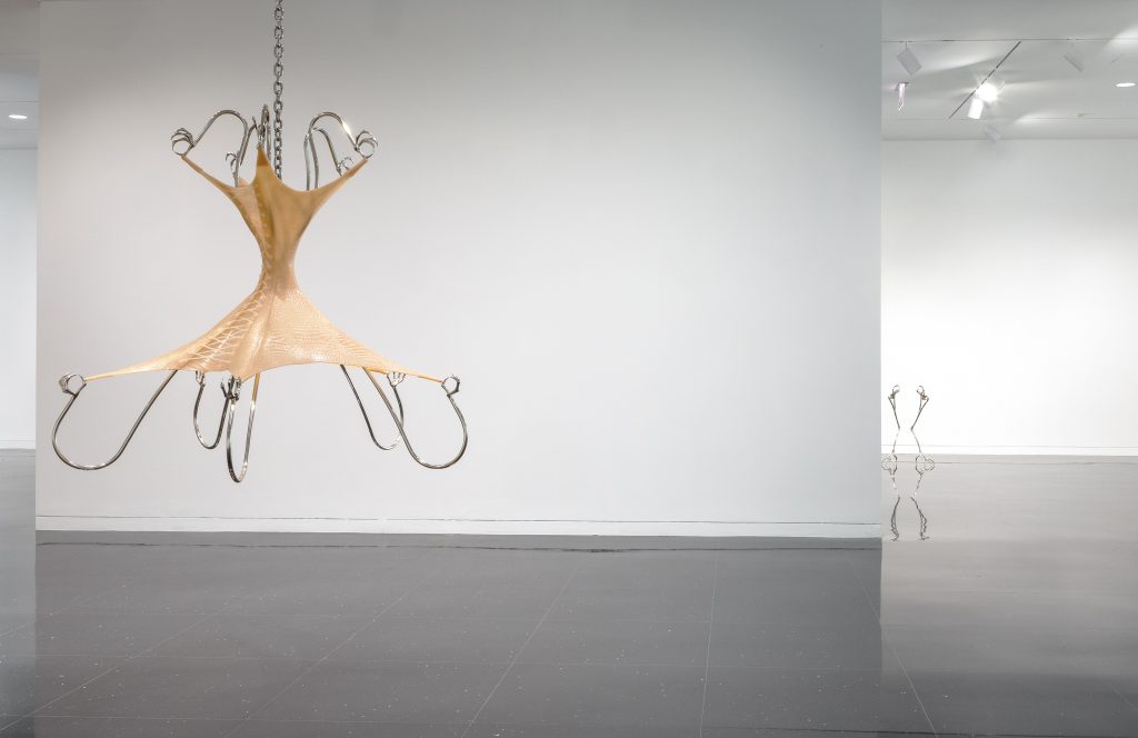 Image: Hannah Levy, "Untitled," 2021. Nickel-plated steel, silicone. 68 x 74 x 74 in. A chandelier-like sculpture that takes the shape of an exaggerated bodice hangs suspended in a white gallery space. The bodice-shape is beige with a corset-like detail. Steel, arm-like, structures appear to be holding the shape in place. Image courtesy of the Arts Club Chicago.