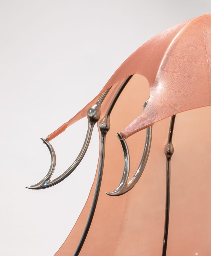 Image: A detail view of Hannah Levy's sculpture "Untitled," 2021. Nickel-plated steel, silicone. 60 x 70 x 85 in. The piece is a crinoline-like, salmon pink sculpture with steel legs. This detail shot shows the points on the steels 'arms' up close, which hold the silicone dome part in place.  Image courtesy of the Arts Club Chicago.