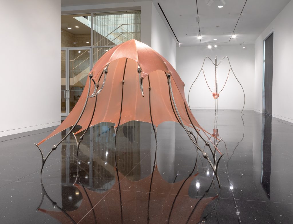 Image: An installation view of Hannah Levy's sculpture "Untitled," 2021. Nickel-plated steel, silicone. 60 x 70 x 85 in. A crinoline-like, salmon pink sculpture with steel legs. The legs appear to be stretching the dome-shape silicone and holding it in place while balancing on the points of its legs. Behind the sculpture to the right is another piece by Levy: Untitled, 2018. Nickel-plated steel, silicone, rubber, zipper. 105 x 97 x 97 in. Rennie Collection, Vancouver. Image courtesy of the Arts Club Chicago.