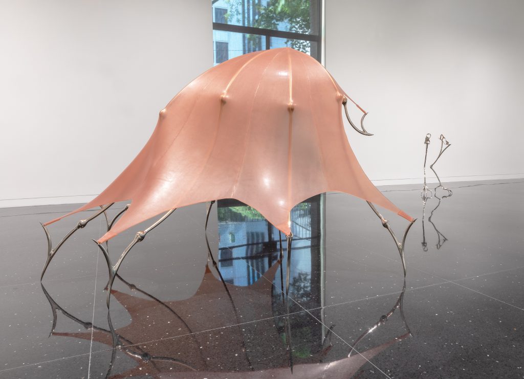 Image: Hannah Levy, "Untitled," 2021. Nickel-plated steel, silicone. 60 x 70 x 85 in. A crinoline-like, salmon pink sculpture with steel legs. The legs appear to be stretching the dome-shape silicone and holding it in place while balancing on the points of its legs. Image courtesy of the Arts Club Chicago.