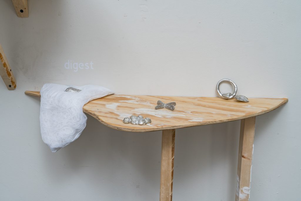 Image: Installation view of UTENSIL at Comfort Station. Four small pewter works ed by Maggie Wong and created by Maggie Wong and Alden Burke are displayed on a curved, wooden shelf. One piece sits on white fabric and has the word "digest" above it on vinyl on the wall. Image courtesy of Annas.