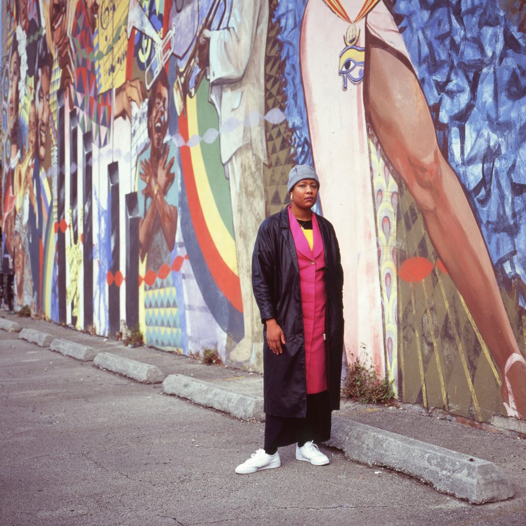 Featured image: A full-length portrait of artist Alkebuluan Merriweather. She is standing and is wearing a gray beanie, a pink dress and a black leather trench coat with white sneakers. Behind her is a mural at the west wall of the Regal Theater displaying images of artists including Billie Holiday, Stevie Wonder, Nat King Cole, and Josephine Baker among others. Along the mural walls are wheel stops made of precast concrete. Photo by Samantha Cabrera Friend.