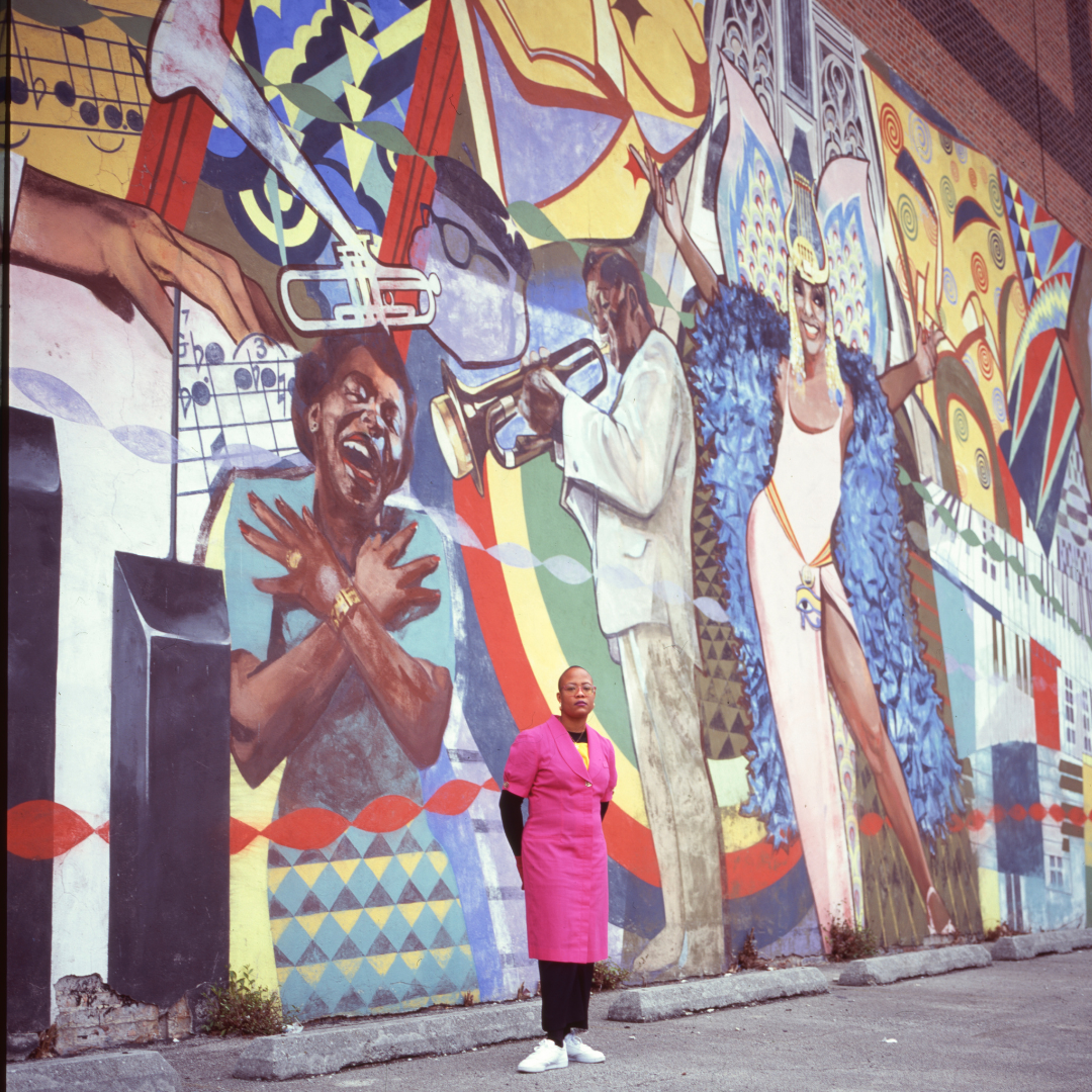 Image: A portrait of artist Alkebuluan Merriweather. She is standing in front of a mural and is  wearing a pink dress and black undergarments of a long sleeve black shirt and pants with white sneakers. The mural behind her is at the west wall of the Regal Theater displaying images of artists including Billie Holiday, Stevie Wonder, Nat King Cole,  and Josephine Baker among others. Along the mural walls are wheel stops made of precast concrete. Photo by Samantha Cabrera Friend.