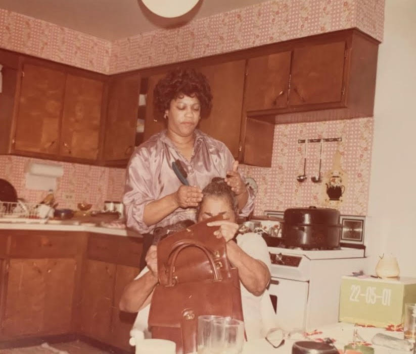 Image: A photograph of Beverly Coleman styling Ora Coleman's (Mama Bede) edges in a kitchen. Beverly has a brush in her hand and is standing behind Ora, who is sitting with a brown leather bag on her lap. Image submitted to @homagetoblkmadonnas by Andrea Coleman. Image courtesy of @homagetoblkmadonnas.