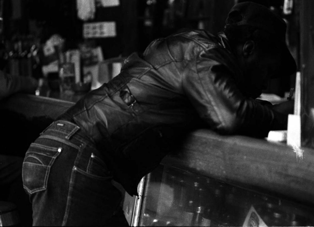 Image: A horizontal black and white photograph of a Black man standing at the bar, writing. He’s wearing a leather jacket, denim pants and a paperboy hat. Underneath the bar is a slightly visible array of bottled beers. The background is less focused, however there’s another patron’s arm in the photo. [Patric McCoy: “Dear Mama,…” a brother writing a letter inside the Rialto at the bar.] Photo taken by Patric McCoy, viewable at the courtesy of the Patric McCoy archive.