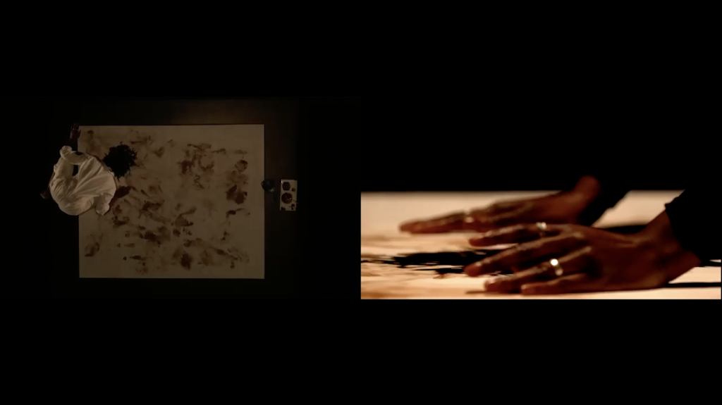 Image: A screenshot of Evidence, divided into two frames. The camera on the left is from directly above Akinbola's canvas, showing Akinbola on the left hand side of the frame, wearing a white suit. The canvas has a a variety of brown markings. The right image is up close on Akinbola's hands, with a ring on her finger. Image by Benji Hart.