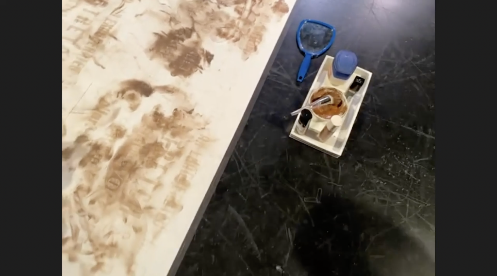 Image: A screenshot taken during Bimbola Akinbola's performance Evidence. A canvas used in the performance is on the left. To its right, in a small white tray on a scratched marley dance floor, are cosmetics in a small white tray. There is a blue-framed hand mirror. Image by Benji Hart.
