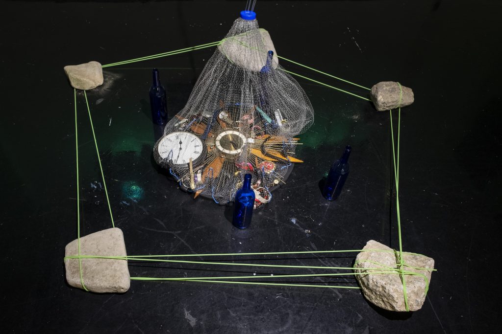 Image: A.J. McClenon, Positive Looping of Integral Retrieval (PLIR) Study, 2021. Casting net, black T-shirts, durags, watches/clocks, fishing cord, rocks, glass bottles, and green lightbulb. Dimensions Vary. Various items tied up in a net in the middle of a hexagon shape made from rocks and green string set on a black floor. Photo by Arda Asena, courtesy of curator Gervais Marsh.