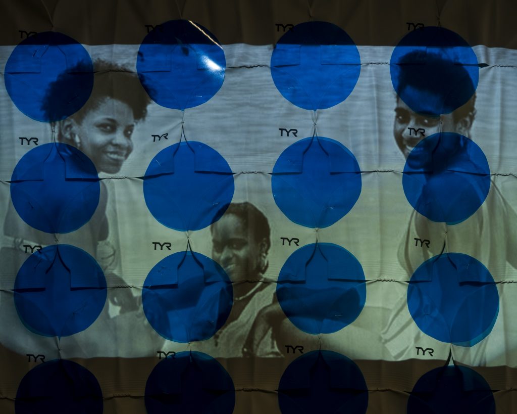 Image: A.J. McClenon, TYR Lake Michigan Studies: All Sides of Water, 2021. Video Projection, swimming caps, cellophane paper. The image is primarily blue and green with poke-dots and three portraits of women. Photo by Arda Asena, courtesy of curator Gervais Marsh.