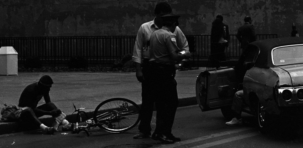 Image: A black and white photograph of a Chicago police officer interacting with a Black man (centered, foreground); left of them is a Black man sitting on the city curb, looking down, at the ground. A bicycle is also laid in front of the man with its front wheel erect. To the right of the officer and civilian is a convertible car with the driver’s door open; a leg is out of the car, foot planted on the ground. In the background are civilians walking by a black fence in front of a weathered city wall. [Patric McCoy: Bicycle/car accident on Jackson and Dearborn, Chicago Loop. Indicative of Loop as Black neighborhood (the bike culture that we have now didn’t exist then so his presence suggests a comfort ability.] Photo taken by Patric McCoy, viewable at the courtesy of the Patric McCoy archive.