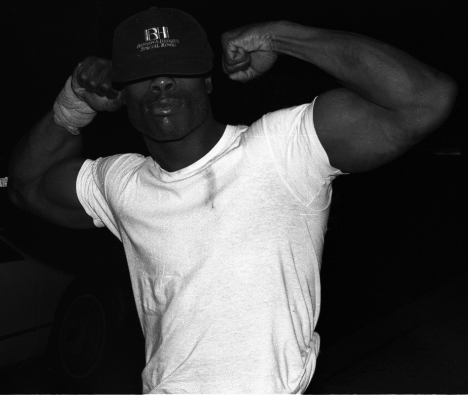 Image: A horizontal, black and white photograph of a Black man wearing a baseball cap that covers his eyes. There’s a stain on the top left part of the t-shirt (right-side in the photo). He’s not smiling, flexing his muscles and seemingly aware that the photo is being taken. There is a sweatband on his right arm. The background of the photo is washed out by the flash of the camera. [Patric McCoy: Strong Man/Tony. Did not go in the Rialto, but for 10 years hustled on State Street and was homeless living on lower Wacker Drive (he was the enforcer down there). He always maintained a membership in the health club so that he could keep clean and pump iron! He didn’t say much and always had his cap pulled down so that you could not see his eyes.] Photo taken by Patric McCoy, viewable at the courtesy of the Patric McCoy archive.