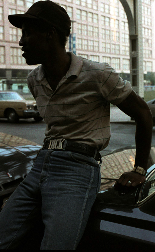 Image: A color photograph of a Black man resting on the top of a car. He’s wearing a neutral colored polo with green stripes, a baseball cap, a bracelet, ring on his wedding finger, blue jeans with a belt buckle with the word, “DICK’; all capital letters. He’s looking away from the camera. Behind him, is a parked car — across the street and support beam of the aboveground L train. In the background is a building that appears to extend for at least half of a city block; it fills the entire background of the photo. [Patric McCoy: Mike sitting on a car outside of the Rialto advertising his wares! He was part of a set of men that just hung around outside of the Rialto but wouldn’t go in. He did frequent the undercover bars in the Chicago Ave. and Clark Street area that catered to the guys coming out of Cabrini Green projects.] Photo taken by Patric McCoy, viewable at the courtesy of the Patric McCoy archive.