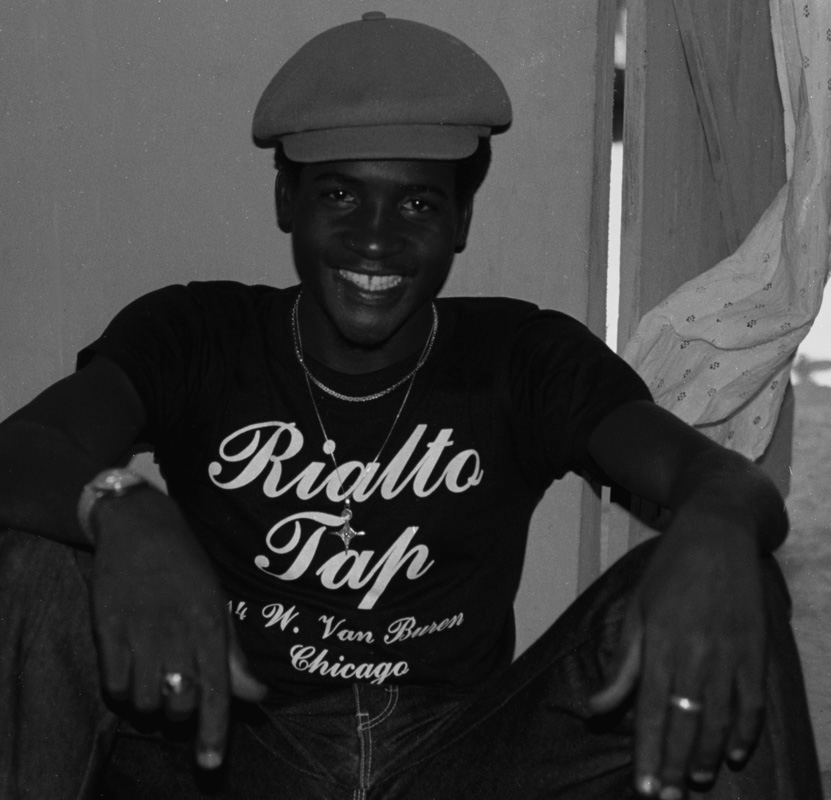 Image: A black and white photograph of a Black man smiling, sitting with his legs open and his hands perched over his knees. He’s wearing a watch on his right arm, a ring on each middle finger, two chains, denim, a t-shirt that says, ‘Rialto Tap (top); 14 W. Van Buren (center) and Chicago (underneath)’, and a paperboy hat. He has a gap between his two front teeth. He is looking directly at the camera. [Patric McCoy: Typical look of trade i.e. masculine presenting but not hyper masculine.] Photo taken by Patric McCoy, viewable at the courtesy of the Patric McCoy archive.