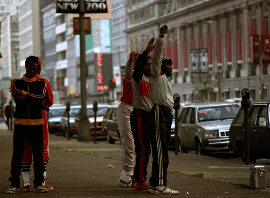 Image:  A photograph of five Black men wearing tracksuits and sneakers. Three of the men are in a line formation with their right hands in the air. One man wears black Adidas track pants with white stripes, a gray sweatshirt with red shoulder detail, and a hat. The man next in formation, is wearing black sweatpants, with two red stripes down the side, red Chuck Taylors with black shoe strings, and a gray sweatshirt with a red panel across the center and with black sleeves. The last man in formation with them wears white sneakers, ruched neutral sweatpants with one small red stripe down the side, and a red sweatshirt with gray sleeves. The three men are facing a street with a row of parked cars. The other two men are to the left of them, standing back to back. The man facing the camera is wearing a black and yellow track jacket with two stripes, black pants and white sneakers. His arms are crossed. The man behind him is wearing an all red tracksuit with the pant legs pulled past his calves that show his white tube socks with three bold black stripes and red low top Chuck Taylors. In the background is a building with pinkish lights that read: ESSEX. The second ‘S’ is the only letter not lit. Other buildings also make up the background of the photo. [Patric McCoy: New Zoo, this was common during that time where guys would perform routines to a boombox on the streets in hopes of being “discovered”. They were performing across from the Conrad Hilton Hotel.] Photo taken by Patric McCoy, viewable at the courtesy of the Patric McCoy archive.