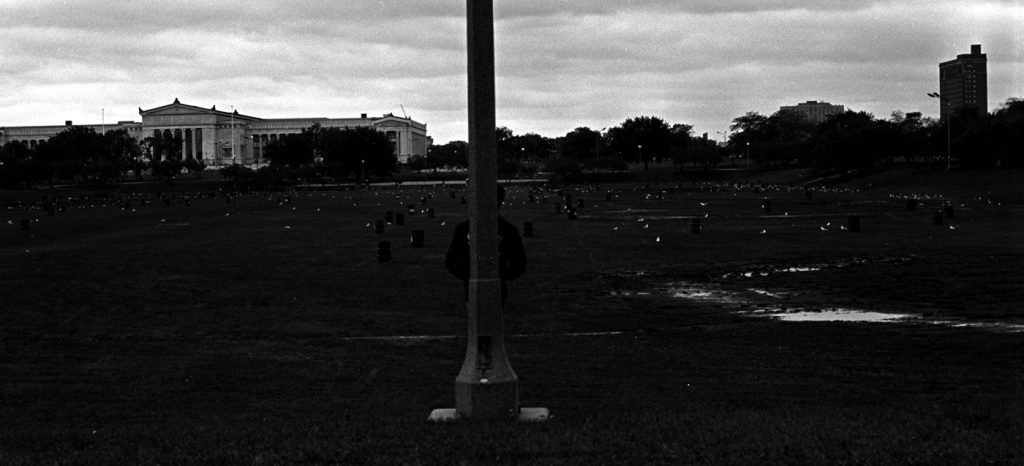 Image: A black and white photograph, taken from Grant Park. In the center of the photograph is a light post, with a figure standing behind it with their back against it. To the left, is the Field Museum. Trees are in front of the museum and span across the entire photograph. There appears to be containers or trash cans scattered across the grass in the park. The sky is bright with very little cloud formation. There’s a tall building to the very far right of photograph in the background. [Patric McCoy: Section of Grant Park long known as a sexual playground and home for the homeless. It was set up for the Bar B Que fest (the garbage cans for the charcoal ashes).] Photo taken by Patric McCoy, viewable at the courtesy of the Patric McCoy archive.