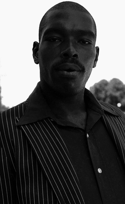 Image: A black and white photograph of a dark-skinned Black man with a thick mustache wearing a pinstripe sports coat and black shirt, with the top buttoned unfastened. The collar of the shirt spills over onto the pinstripes. His hair is in a buzz cut with a crisp lining to frame his angular face. He looks directly into the camera. The background of the photograph is out of focus. [Patric McCoy: Homeless man that frequented the southern part of the downtown area from Jackson to Roosevelt and Grant Park. Always dressed well but would not go into the Rialto.] Photo taken by Patric McCoy, viewable at the courtesy of the Patric McCoy archive.
