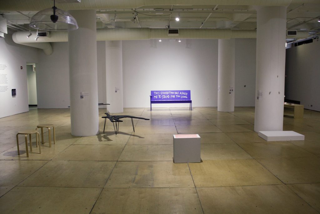 Image: An installation shot of the exhibition *Crip at Gallery 400. Various sculptures are displayed in the gallery. In the back of the gallery, there is a blue bench with text that reads: "THIS EXHIBITION HAS ASKED ME TO STAND FOR TOO LONG". Courtesy of the gallery.