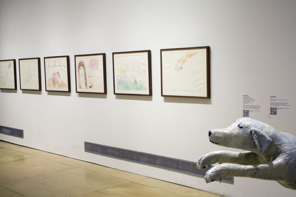 Image: An installation shot of six framed drawings hanging on a white wall by Emilie Gossiaux. The drawings are made with ballpoint pen and crayon on paper and are 17 x 23" each. There is a sculpture of a dog peeking out of the bottom right corner of the frame. The sculpture is titled Dancing with London by Emilie Gossiaux, 2017, aluminum, foam, paper- mache, rubber, resin, and nail polish, 53 x 48 x 18". Courtesy of the artist and Gallery 400.