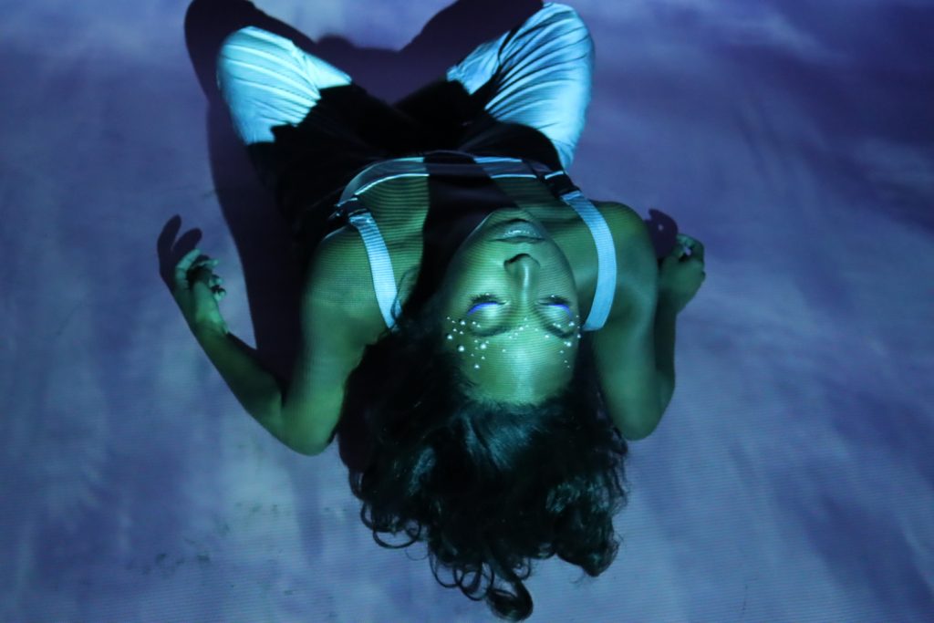 Image: A still from God is a Black Woman by Chaz Shermil Hodges. A Black woman (actress Kalyn Rivers) bends backwards so that her face appears upside down to the viewer. The floor she is on is blue and there is a blue tint to the entire image, giving it an underwater feel. Image courtesy of the author. Photo by Salem Daniel.
