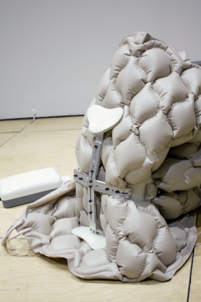 Image: Berenice Olmedo, áskesis, alternating pressure pad, taylor back brace, 2019, arduino boards (BOM/S 1203), 40 x 40 x 83 cm. A beige/gray sculpture made from an alternating pressure pad and other materials is installed in a gallery. Courtesy of the artist and Jan Kaps, Cologne.