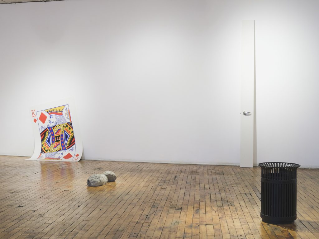 Image: An installation view of General Objects at Heaven Gallery. The four pieces shown are as follows from left or right: Big Boy (Diamonds) by Conrad Cheung [a large king of diamonds card leaning against the gallery wall], (carhartt hat arch) by Gary LaPointe Jr. [two concrete hats facing each other sitting on the gallery floor], Off White and Hickory (Door) by Conrad Cheung [a sliver of a white door with a silver handle leaning against the gallery wall], and Twice as Far Away (Street Trash Can) by Catherine Hu [a life-size black trashcan installed in the gallery]. Image courtesy of Heaven Gallery.