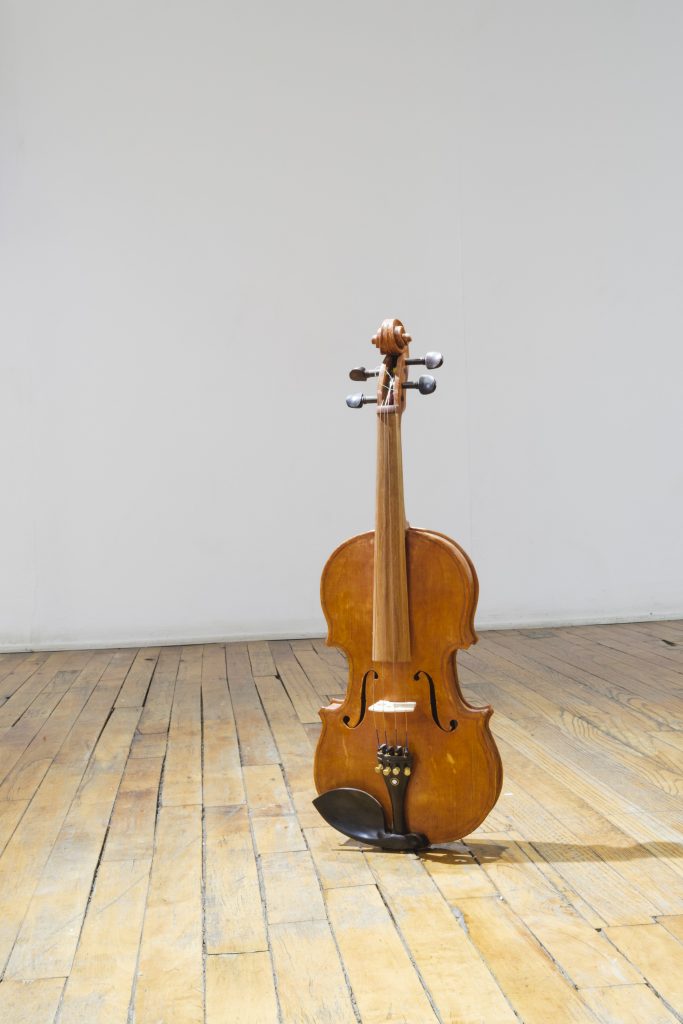 Image: Catherine Hu, Violin, 2019, pine, poplar, cherry, violin hardware, 24" x 8" x 2". A life-sized and realistic-looking violin sits upright on a gallery floor. Image courtesy of Heaven Gallery.