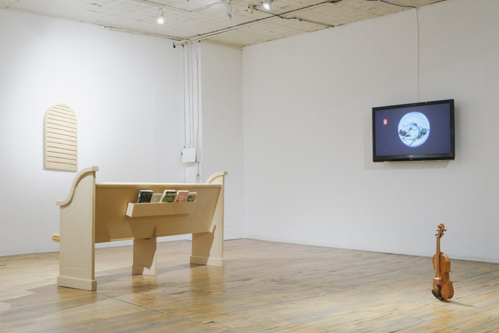 Image: An installation view of General Objects at Heaven Gallery. The four pieces shown are as follows from left or right: Louver and Pew by Clayton Phillips (Wooden slats hanging on a wall and a wooden church-like pew installed in the gallery), white vegetable i by stephanie mei huang (a video on display on a screen on the wall), and Violin by Catherine Hu (a realistic-looking violin sitting upright on the gallery floor). Image courtesy of Heaven Gallery.