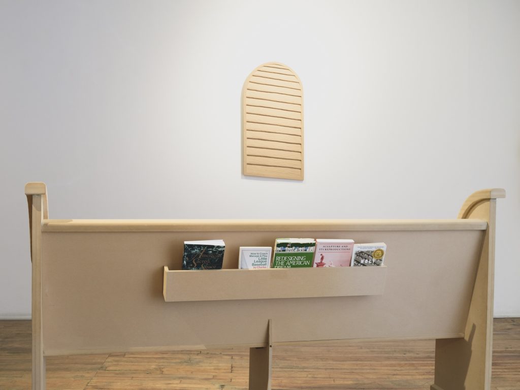 Image: Clayton Phillips, Pew, 2019, MDF, foam, 6' 3" x 16" x 40". Clayton Phillips, Louver, 2020, MDF, 18" x 36". Wooden slats hangs on a wall in the background and a wooden church-like pew is installed in the foreground. Image courtesy of Heaven Gallery.
