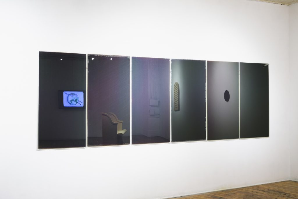 Image: Gary LaPointe Jr., (idle engine, collection of 6 black solar panels), 2019, active solar panels that were previously owned by a family of doomsday preppers, electrical, and hardwire, 151" x 49". Six black solar panels are installed on a white gallery wall. Other artwork in the gallery can be seen reflected in the solar panels. Image courtesy of Heaven Gallery.