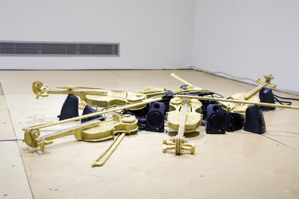 Image: Christopher Robert Jones, PureImagination_Sextet, 2020, OSB, wood glue, twine, USB drive, media players, computer speakers, violin-vocal rendition, 16 x 72 x 48". A sculpture that looks like a pile of yellow violins and speakers installed on the floor of a gallery. Courtesy of the artist.