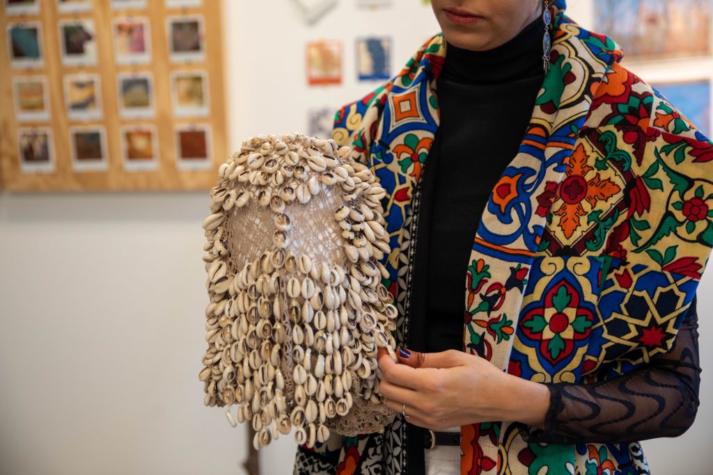 Image: Farah Salem can be seen from mid-face down, holding a headpiece instrument that's covered in cowry shells. You can see her vibrant scarf draped over a black turtleneck top. Part of a wood panel with Polaroids from the 'Erode Re-Compose' hanging in the background. Photo by EdVetté Wilson Jones.