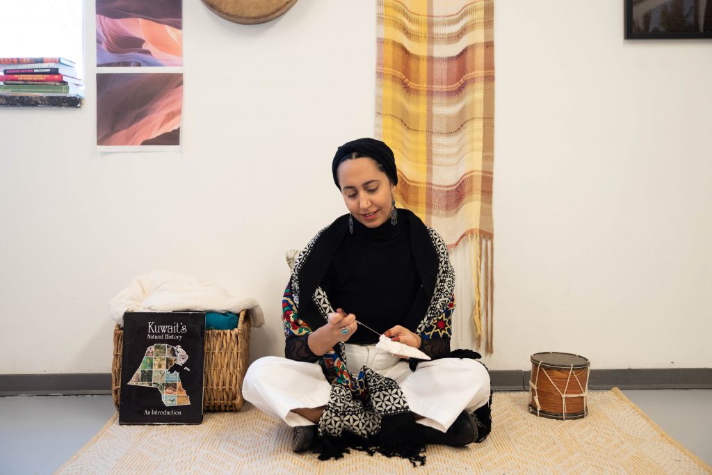 Image: A black-and-white photo of Farah Salem as she sits next to a window on a mat in her studio at Hyde Park Art Center. There are art works, fabrics, instruments, and photographs hanging on the wall behind and above her. Farah is looking down at her hands with a book titled 'Kuwait's Natural History: An Introduction.' Photo by EdVetté Wilson Jones.