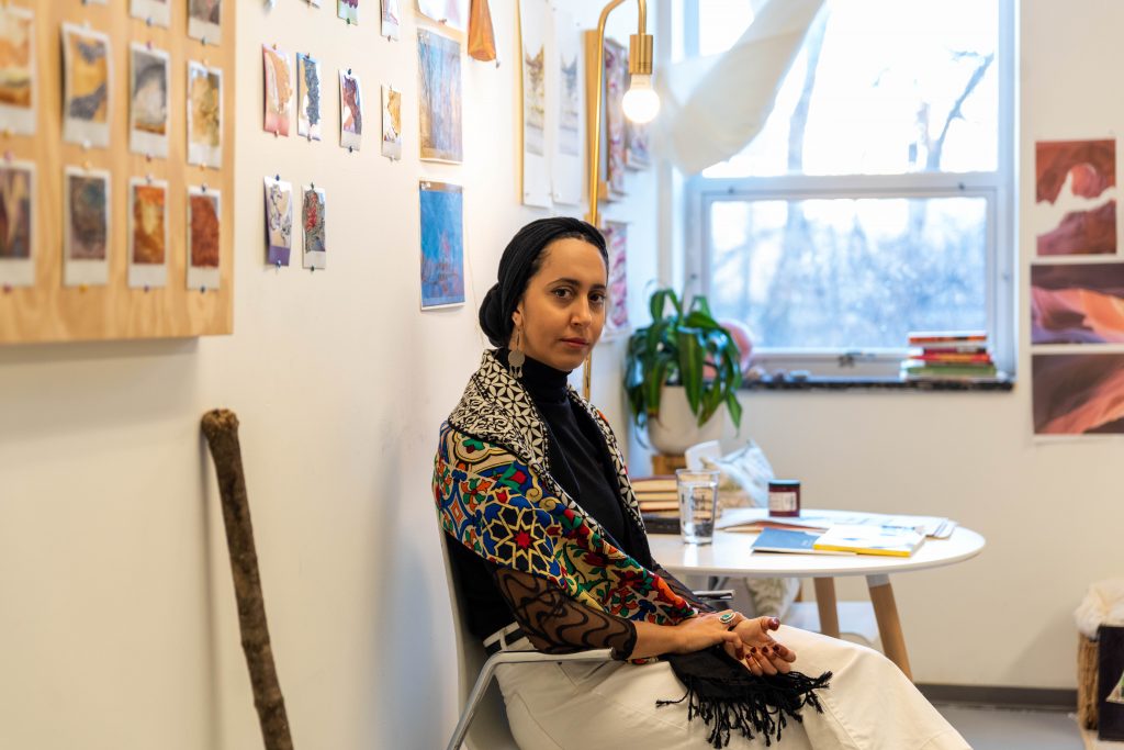 Image: A portrait of Farah Salem sitting in her studio wearing white pants and a vibrant scarf draped over a black turtleneck top. Part of a wood panel with Polaroids from the 'Erode Re-Compose' hanging in the background, with other polaroids directly on the wall. Partial views of images from the series Temporary Deformations. Photo by EdVetté Wilson Jones.