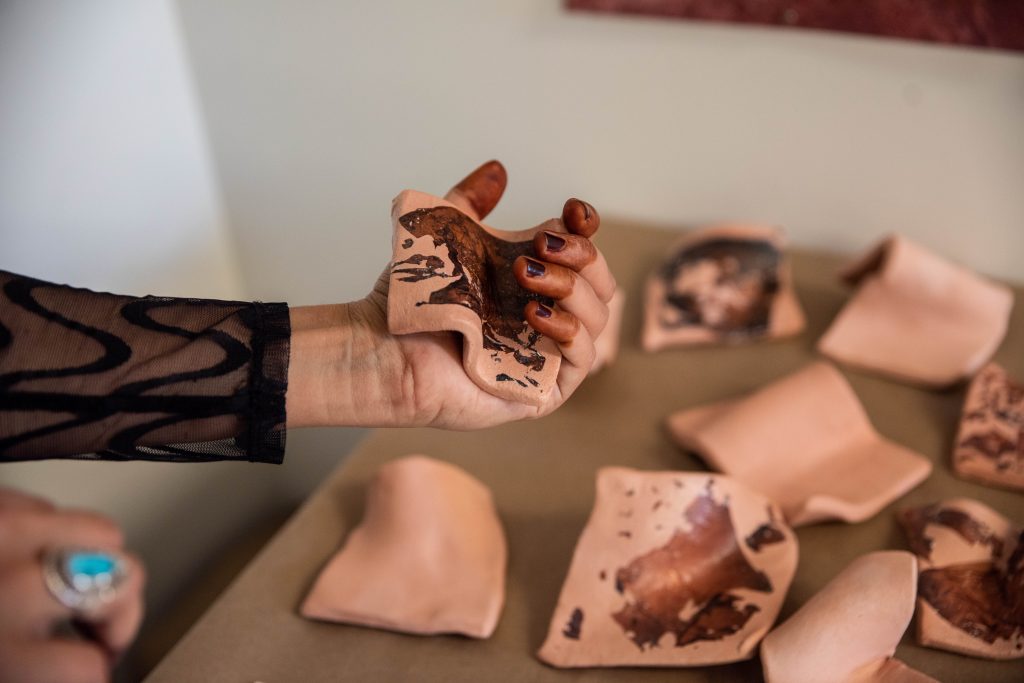 Image: With dyed fingertips and a small grip-shaped sculpture being softly clutched, Farah's hand stretches into the frame from the left. Just beyond her hand you can see more small bright copper-colored sculptures from the series sitting on a table. Photo by EdVetté Wilson Jones.