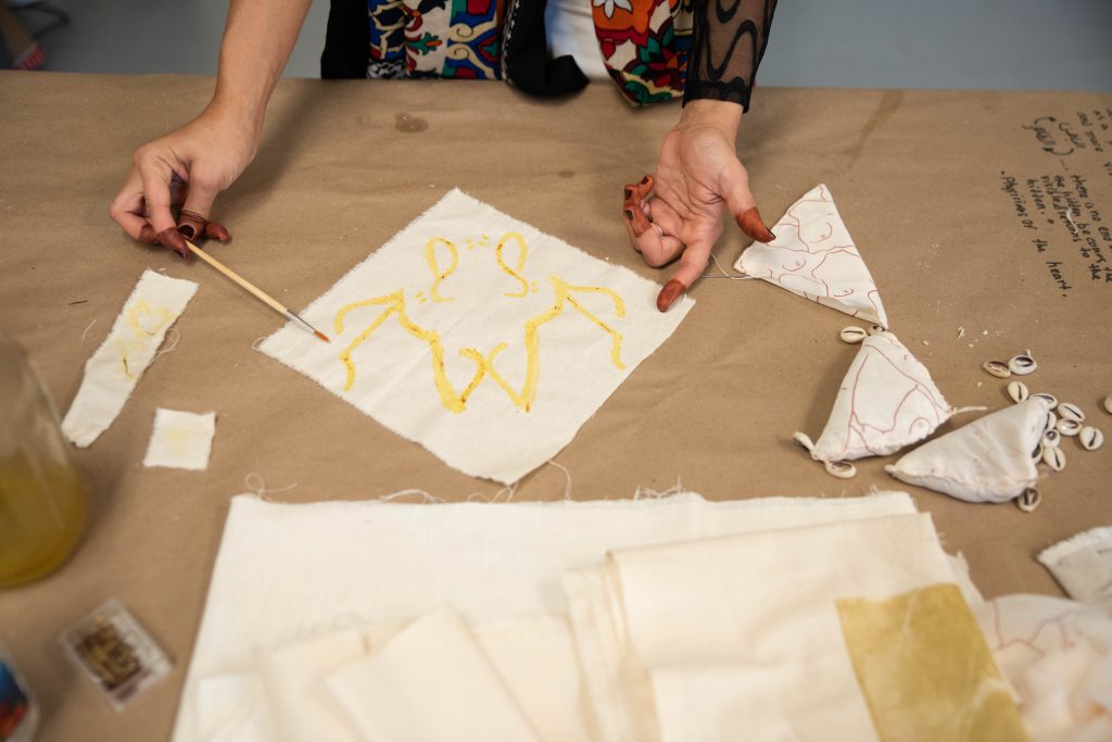 Image: Small pieces of muslin fabric sit folded and stacked on a table covered in butcher paper. You can see Farah's hands at the top of the image, one holding a small paint brush and the other pointing to words "freedom" painted in yellow, mirrored, on the fabric. Photo by EdVetté Wilson Jones.