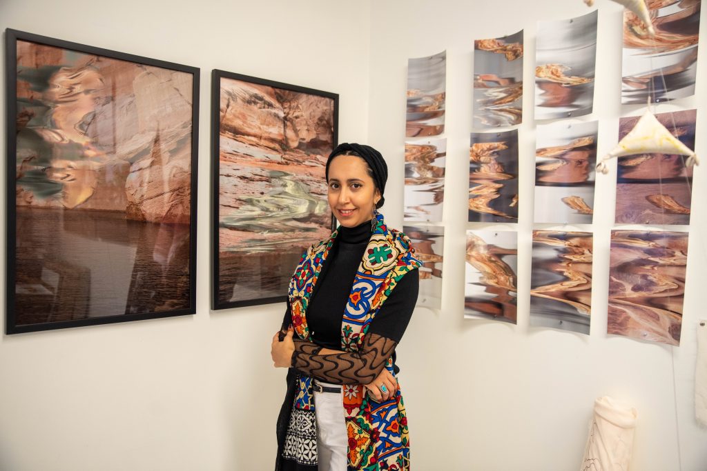 Image: A waist-up portrait of Farah Salem, standing in front of printed images from Temporary Deformations, a series of digital renderings of fiber glitches within desert rock landscapes. Photo by EdVetté Wilson Jones.