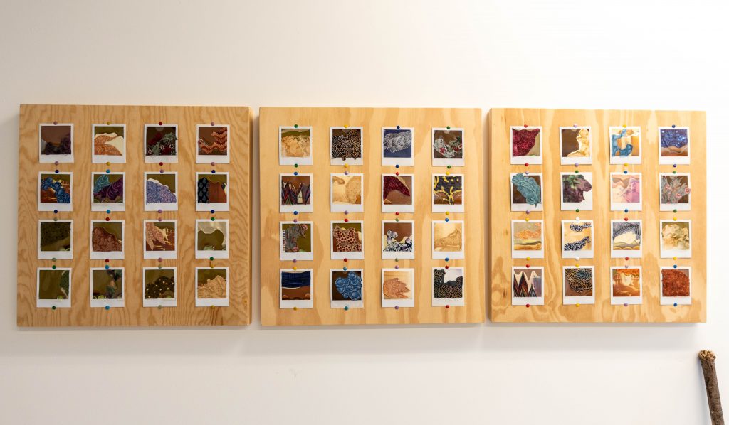 Image: Three square wood panels hang side-by-side on a wall. Each panel has a four by four grid of Polaroid collages mounted to them with colorful thumb tacks, all part of the piece 'Erode Re-Compose.' Photo by EdVetté Wilson Jones.